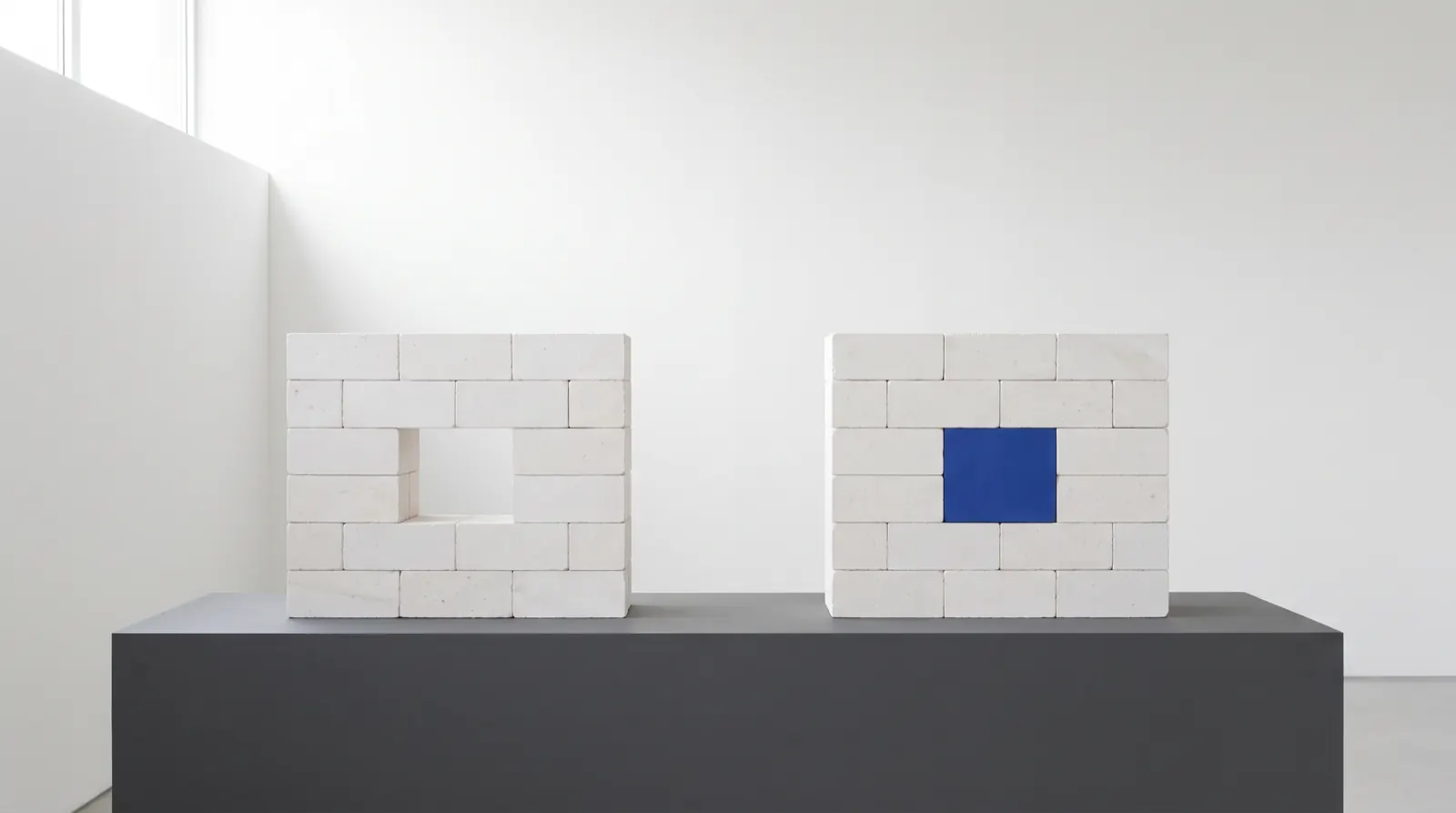 Two identical white block walls on a graphite plinth. The left wall has one block missing; the right wall has the same gap filled with a single blue block.